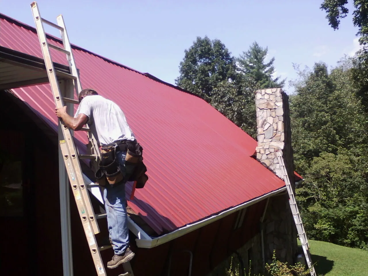 Roofer on a ladder beside a red metal roof and stone chimney — Soffit & Fascia Repair by Roofing Brothers in Thornwood